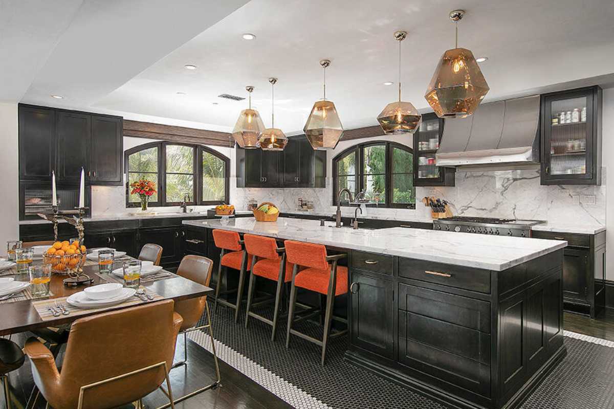 A kitchen with a countertop, a sink, windows, a white ceiling, lighting, cabinets, kitchen appliances, and a table and chairs.