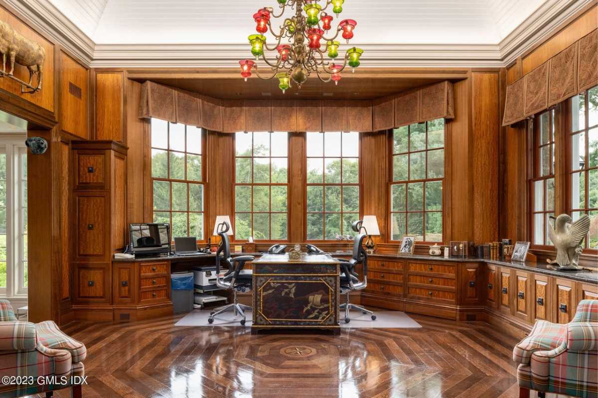 A home office with windows, a chandelier, wooden cabinets, chairs, and a desk.