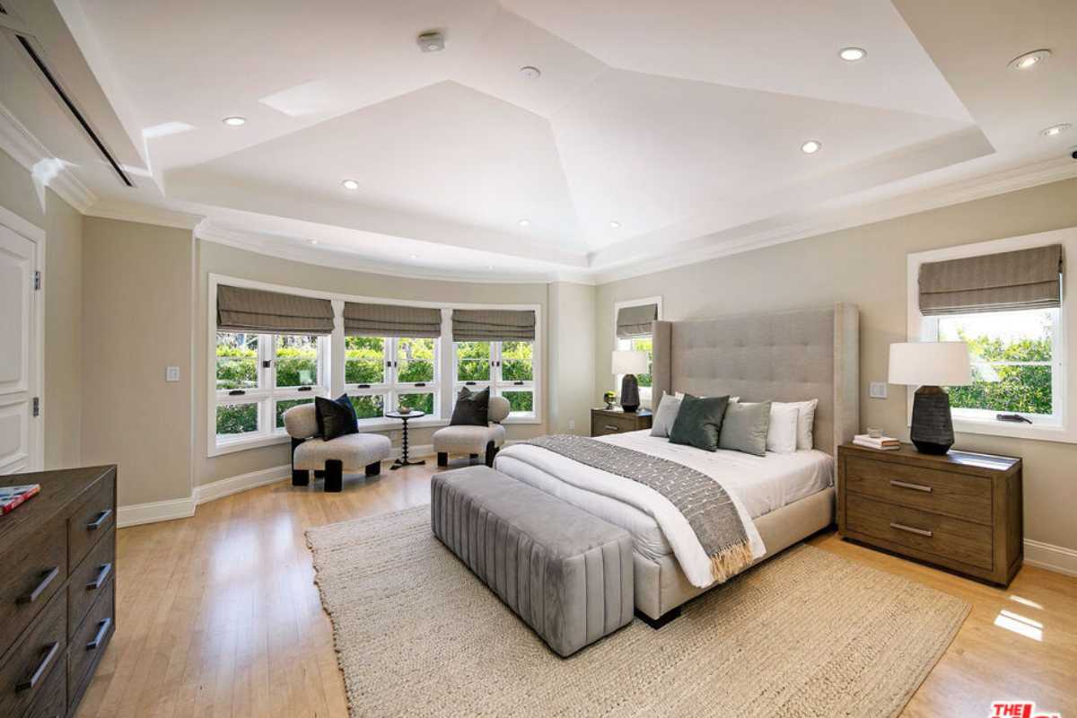 A bedroom with a bed, rug, wooden floor, cabinets, chairs, windows, and a white ceiling.