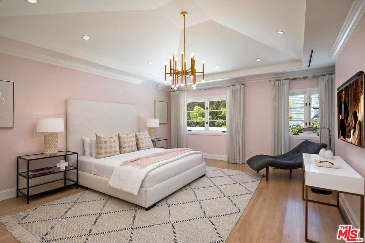 A bedroom with a bed, rug, wooden floor, nightstand, chandelier, chairs, windows, TV, and a white ceiling.