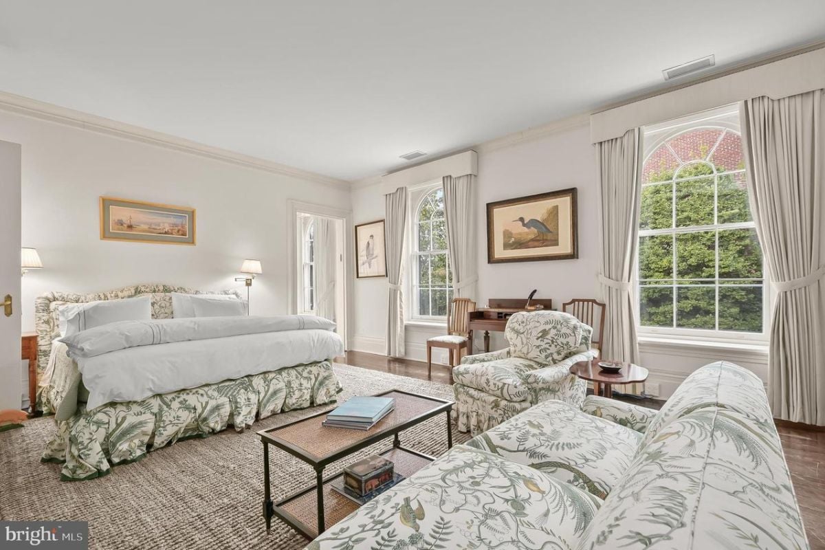 A bedroom with a bed, white ceiling, couches, windows, coffee table, and paintings.