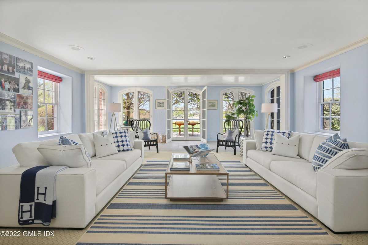 A lounge with a striped rug, white couches, a center table, windows, a white ceiling, chairs, and potted plants.