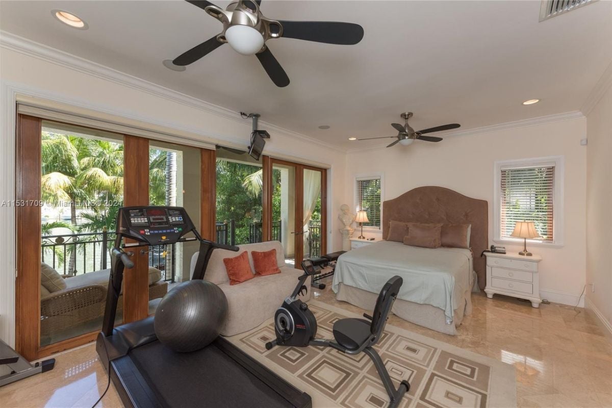 A bedroom with tiled floors, a white ceiling, a ceiling fan, a bed, a nightstand, a lampshade, a rug, and gym equipment.