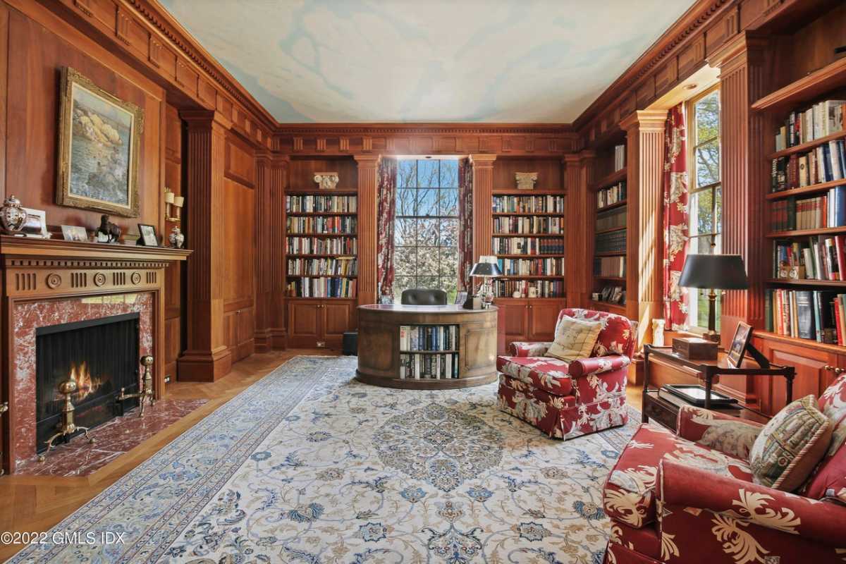 A home office with a fireplace, rug, wooden bookshelves filled with books, chairs, and a desk.