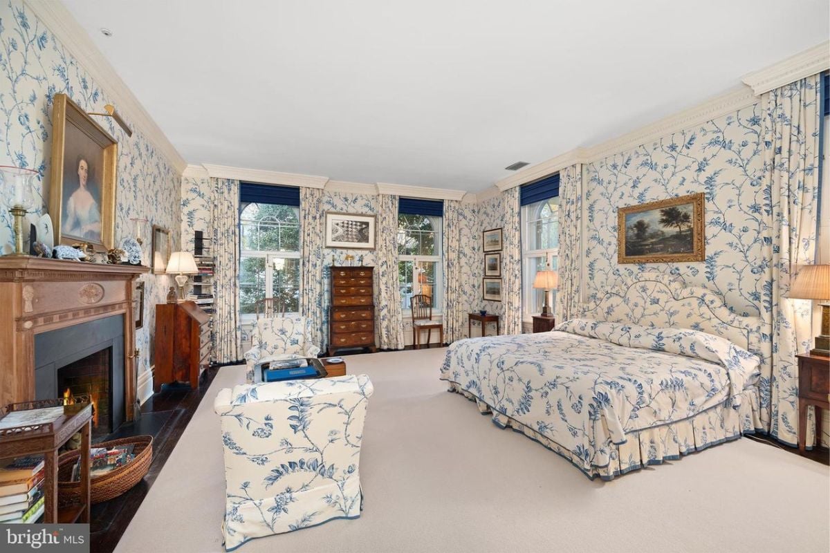 A bedroom with a fireplace, wallpaper, white floor, white ceiling, nightstand, and lampshade.