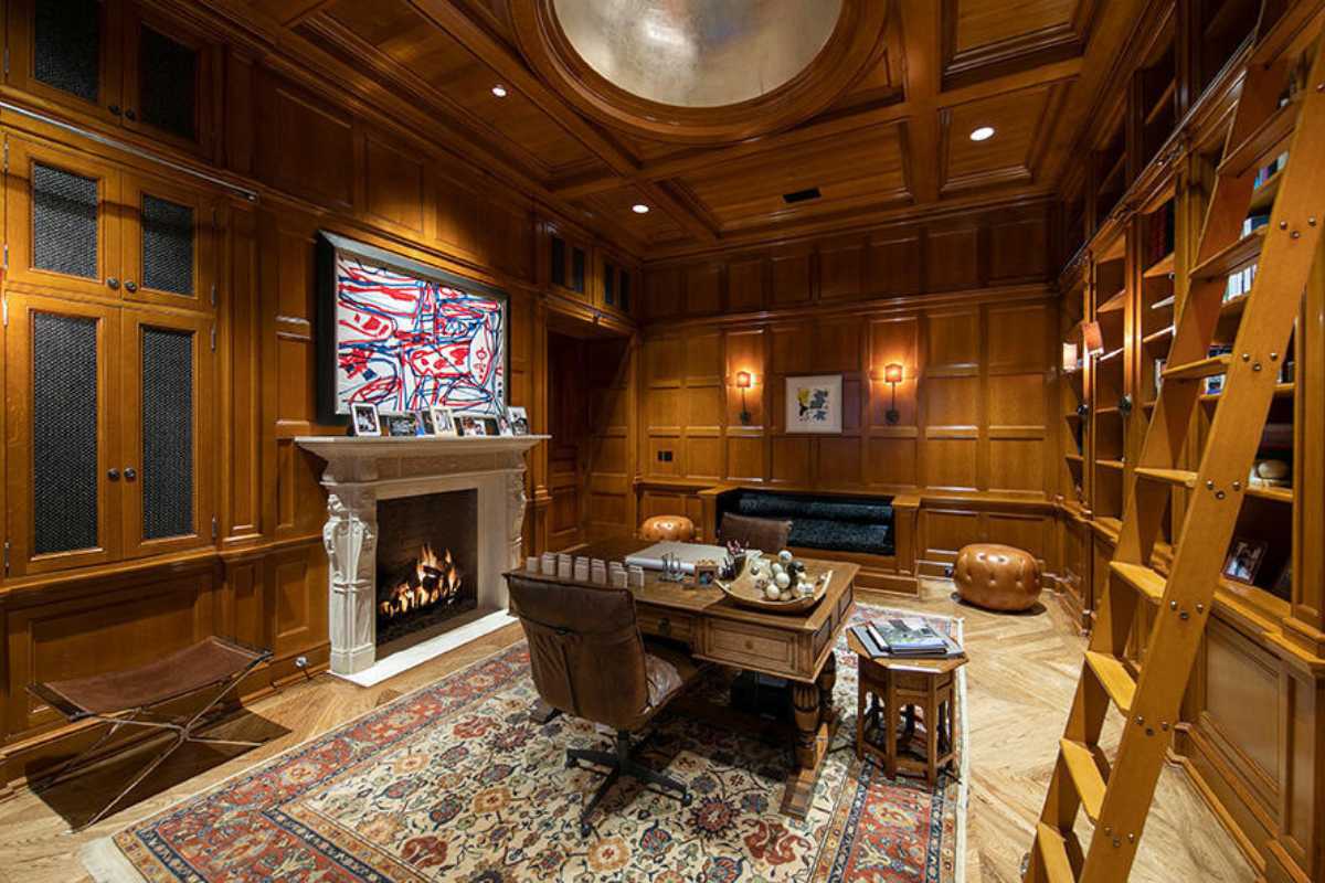 A home office with a rug, a ceiling with molding, a fireplace, a painting, cabinets, shelves, a ladder, a desk, and chairs.