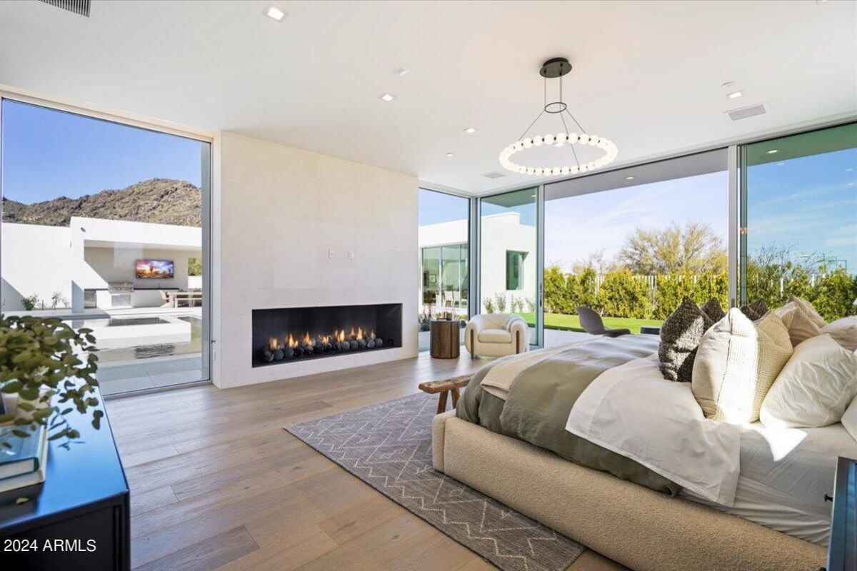 A bedroom with a wooden floor, white ceiling, chandelier, fireplace, bed with pillows, and rug.