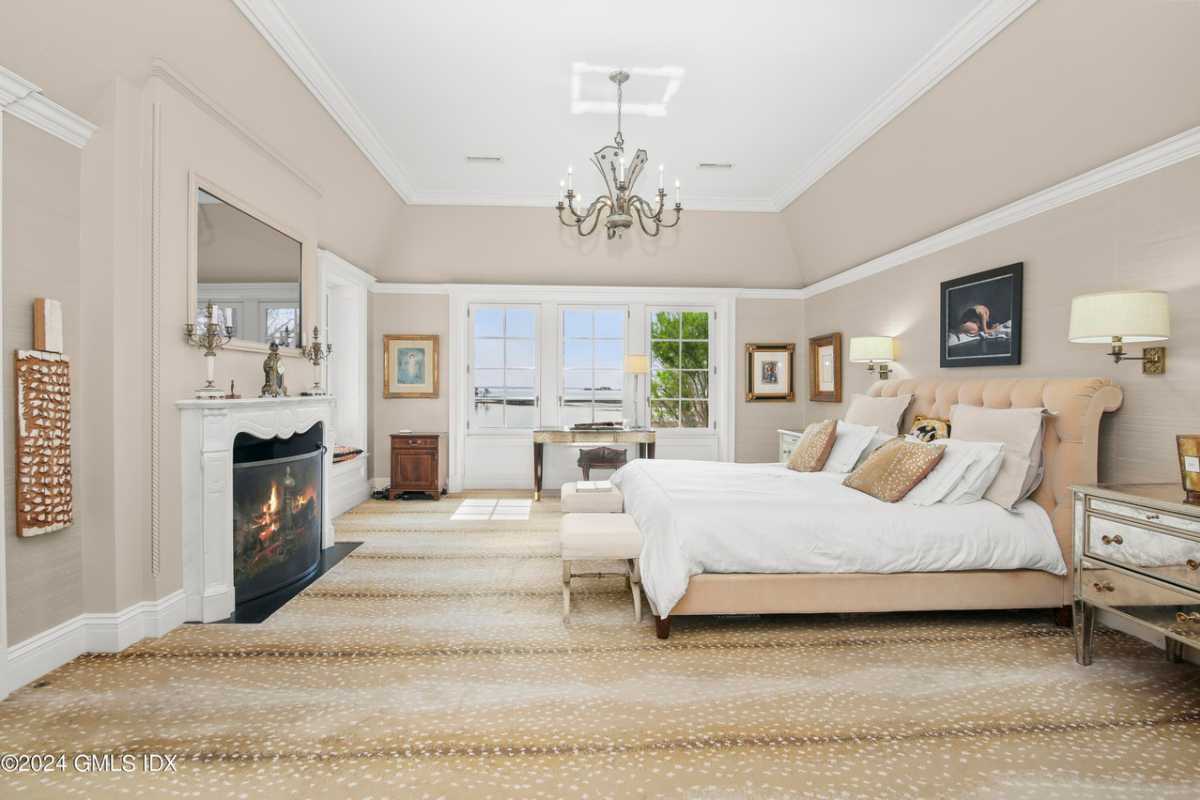 A bedroom with a large bed, unique flooring, a fireplace, a nightstand, a window, a chandelier, and a white ceiling.