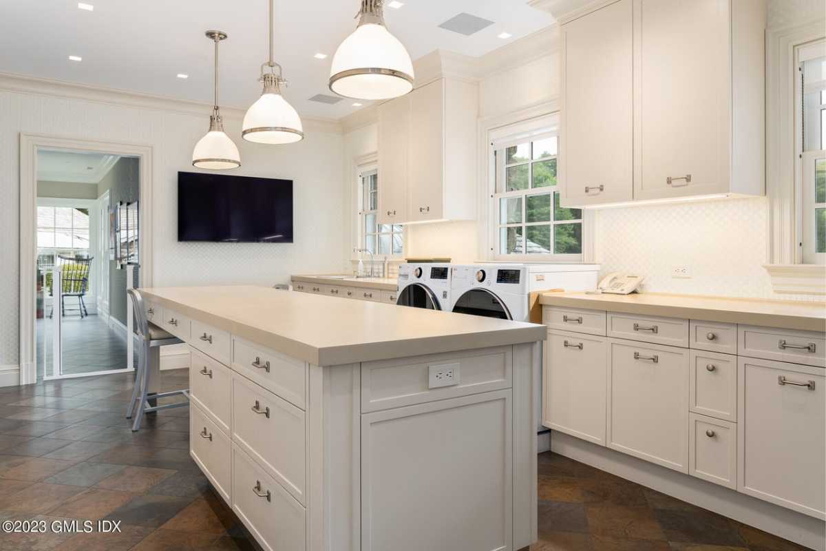 A laundry room with a white island, TV, white ceiling, lighting, windows, washing machine, and white cabinets.