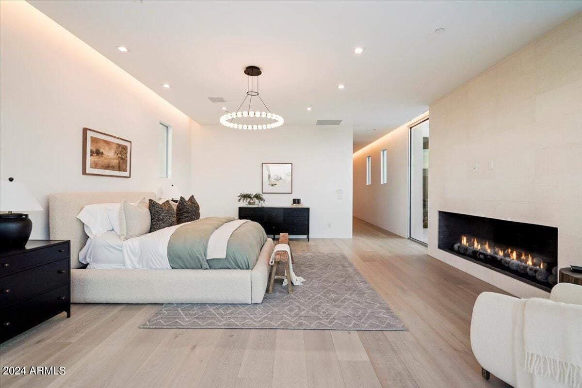 A bedroom with a wooden floor, white ceiling, chandelier, fireplace, bed with pillows, and rug.