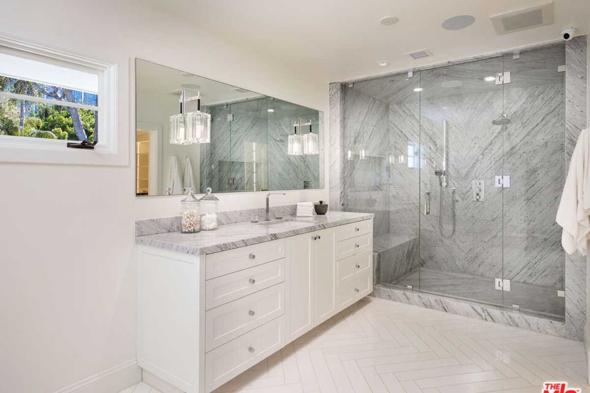 A bathroom with a tiled floor, sink, mirror, white cabinets, and a shower room.