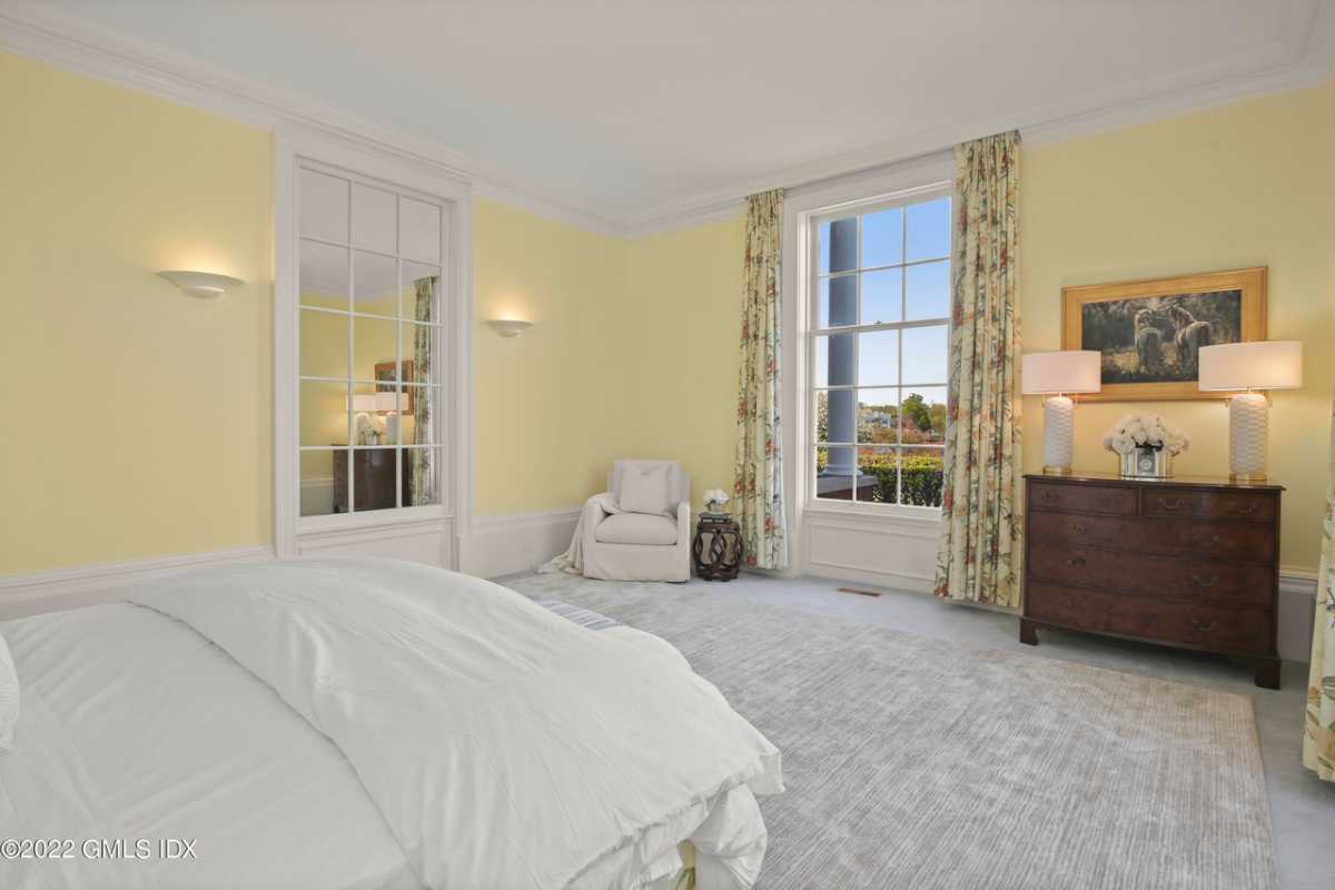 A bedroom with a bed, cabinet, chair, lampshade, windows, white walls, and a white ceiling.