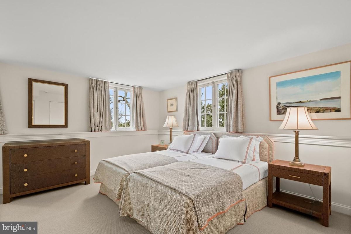 A bedroom with a bed, white floor, white ceiling, nightstand, lampshade, painting, and windows.