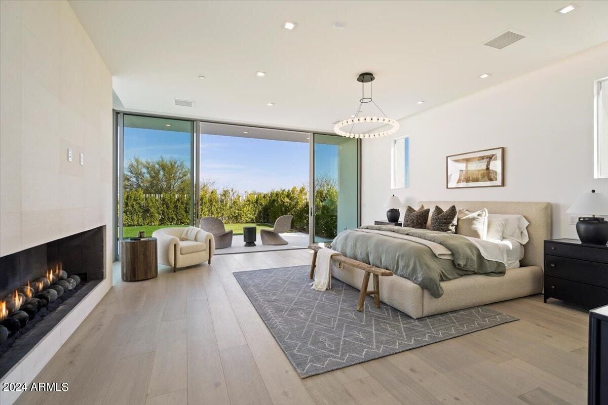 A bedroom with a wooden floor, glass door, white ceiling, chandelier, fireplace, bed with pillows, and rug.
