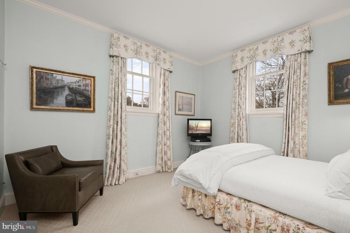 A bedroom with white walls, white ceiling, bed, single couch, TV, and windows with curtains.