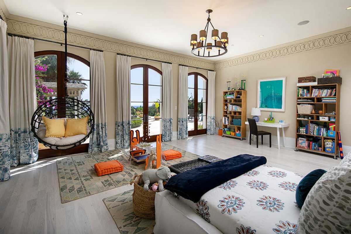 A bedroom with a bed, a rug, tiled floor, a chandelier, a shelf, windows, chairs, and a table.