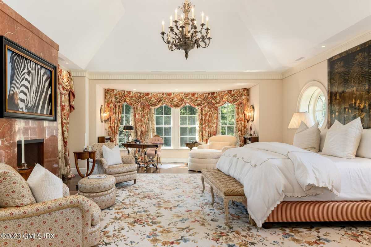 A bedroom with a bed, rug, fireplace, windows, white ceiling, chandelier, painting, and couches.