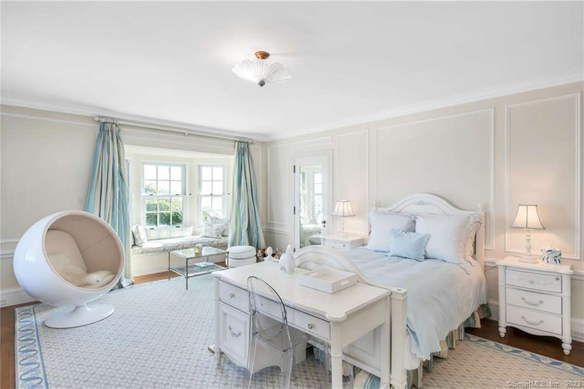 A bedroom with a white bed, a rug, white ceiling, white walls, a window, a chair, a white nightstand, and lampshades.