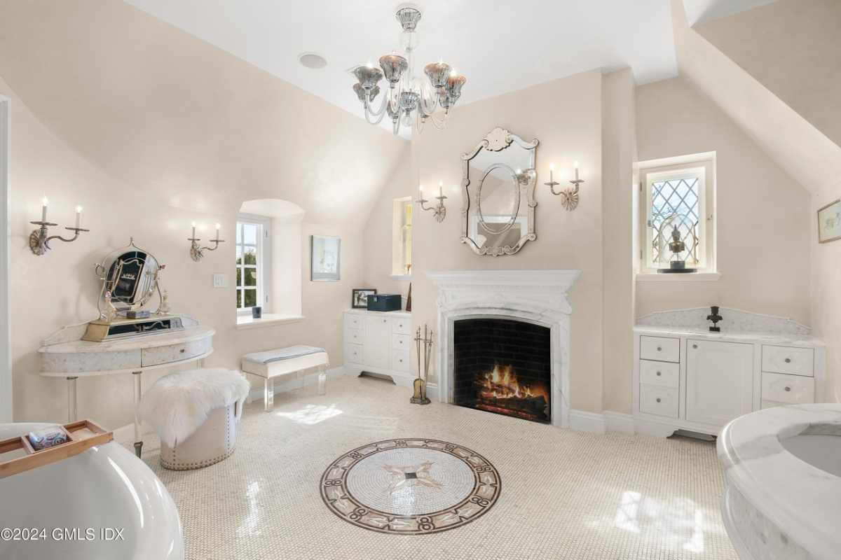 A bathroom with a fireplace, tiled floor, a chandelier, a bathtub, white cabinets, windows, a mirror, a white ceiling, and a chandelier.
