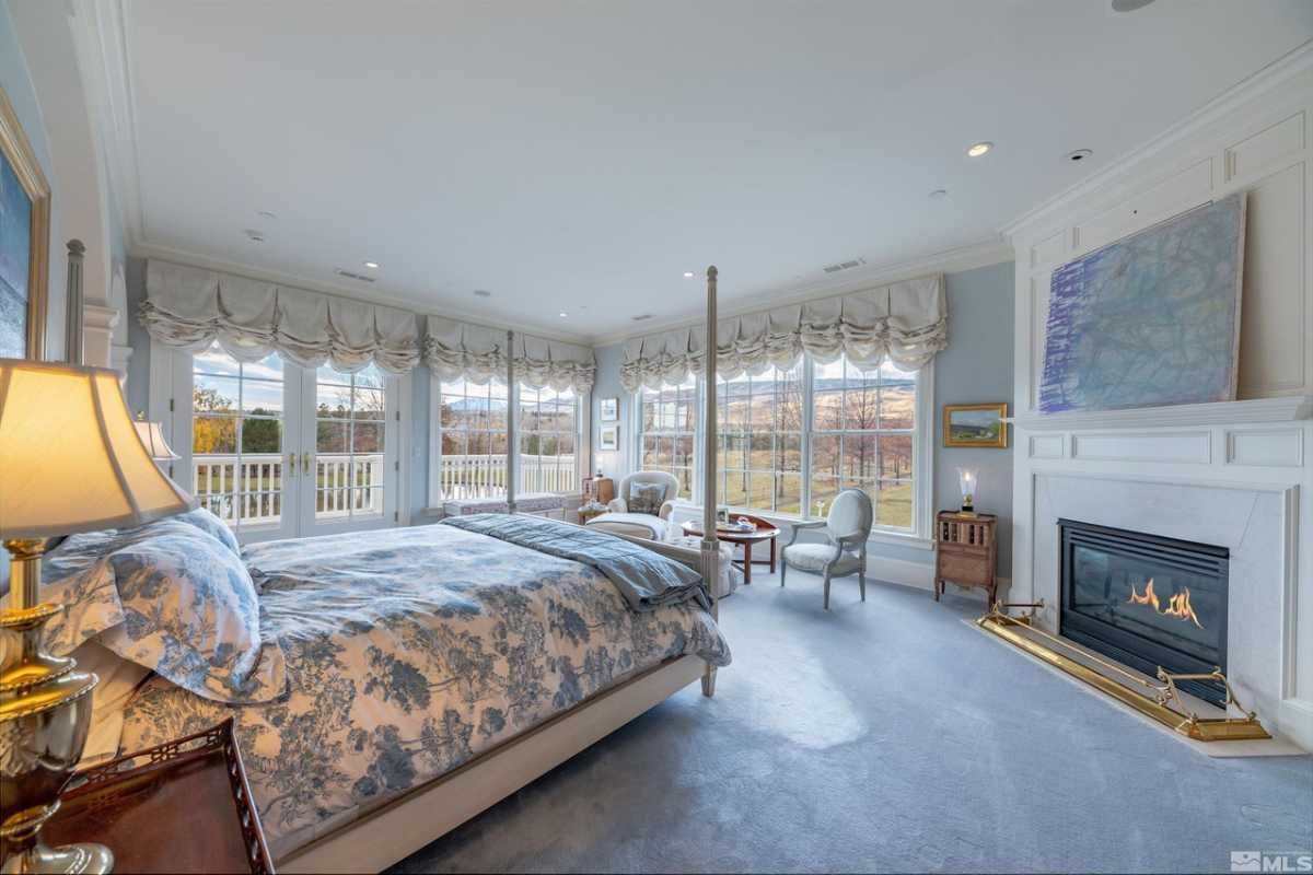 A bedroom with a bed, windows, chairs, a white ceiling, a painting, and a fireplace.
