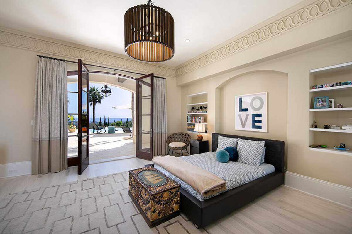 A bedroom with a rug, a bed, shelves, a chair, a white ceiling, and a chandelier.