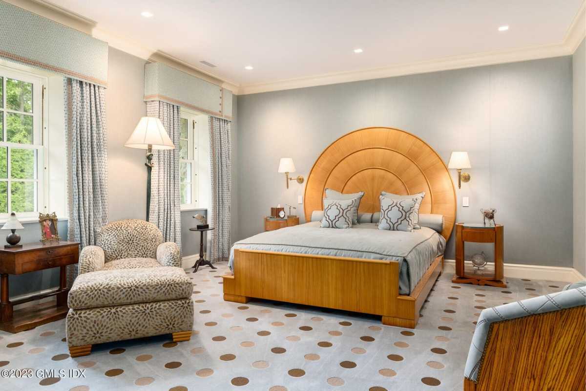 A bedroom with a bed, nightstand, lampshade, gray walls, windows, white ceiling, and a couch.