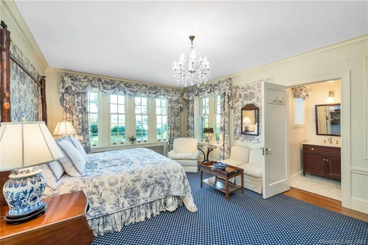 A bedroom with a bed, a rug, a nightstand, lampshades, windows with curtains, a white ceiling, and a chandelier.