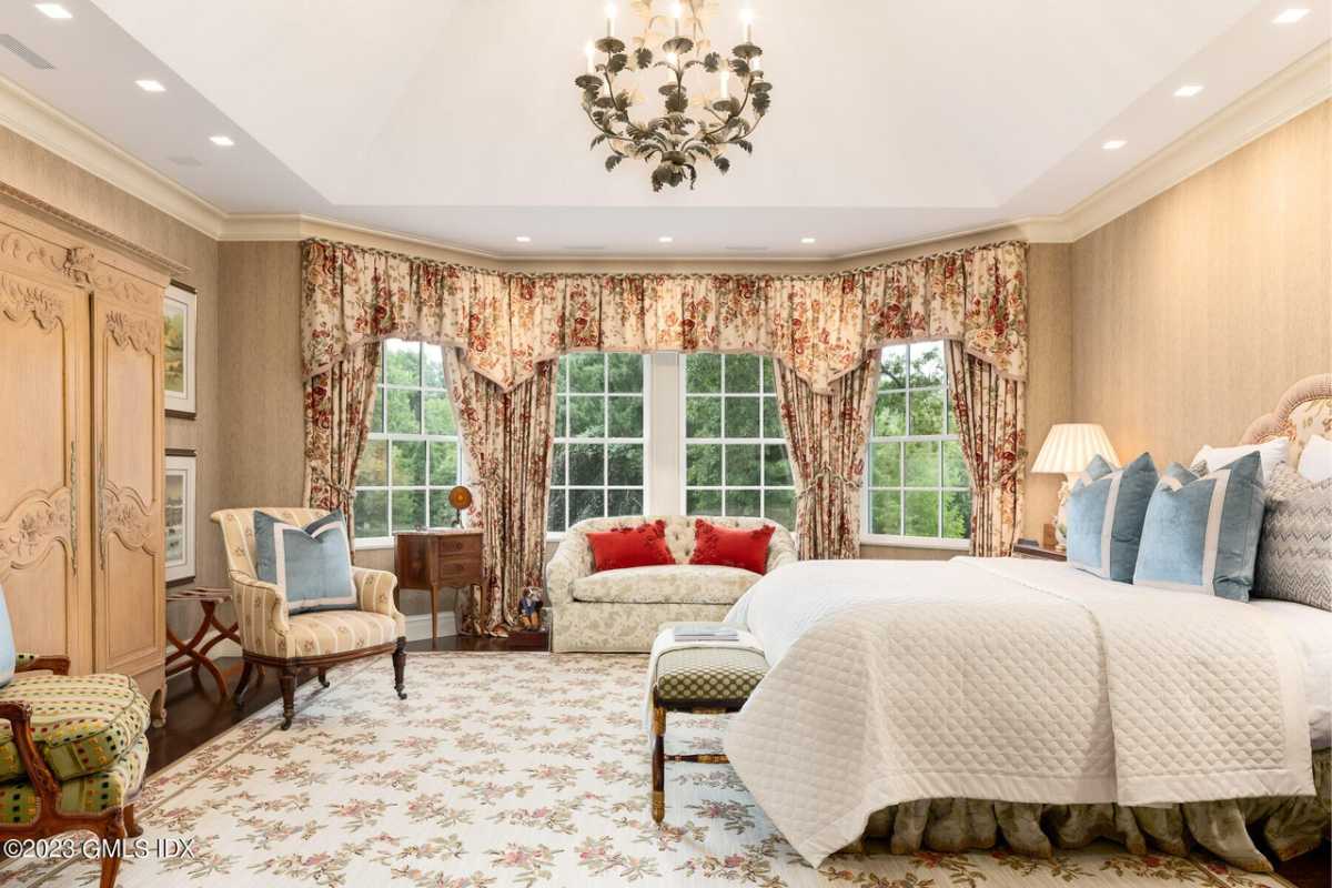 A bedroom with a bed, rug, windows, white ceiling, chandelier, cabinets, couch, and chairs.