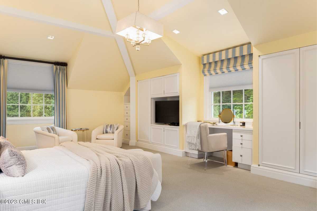 A bedroom with a bed, yellow walls and ceiling, TV, windows, cabinets, and chairs.