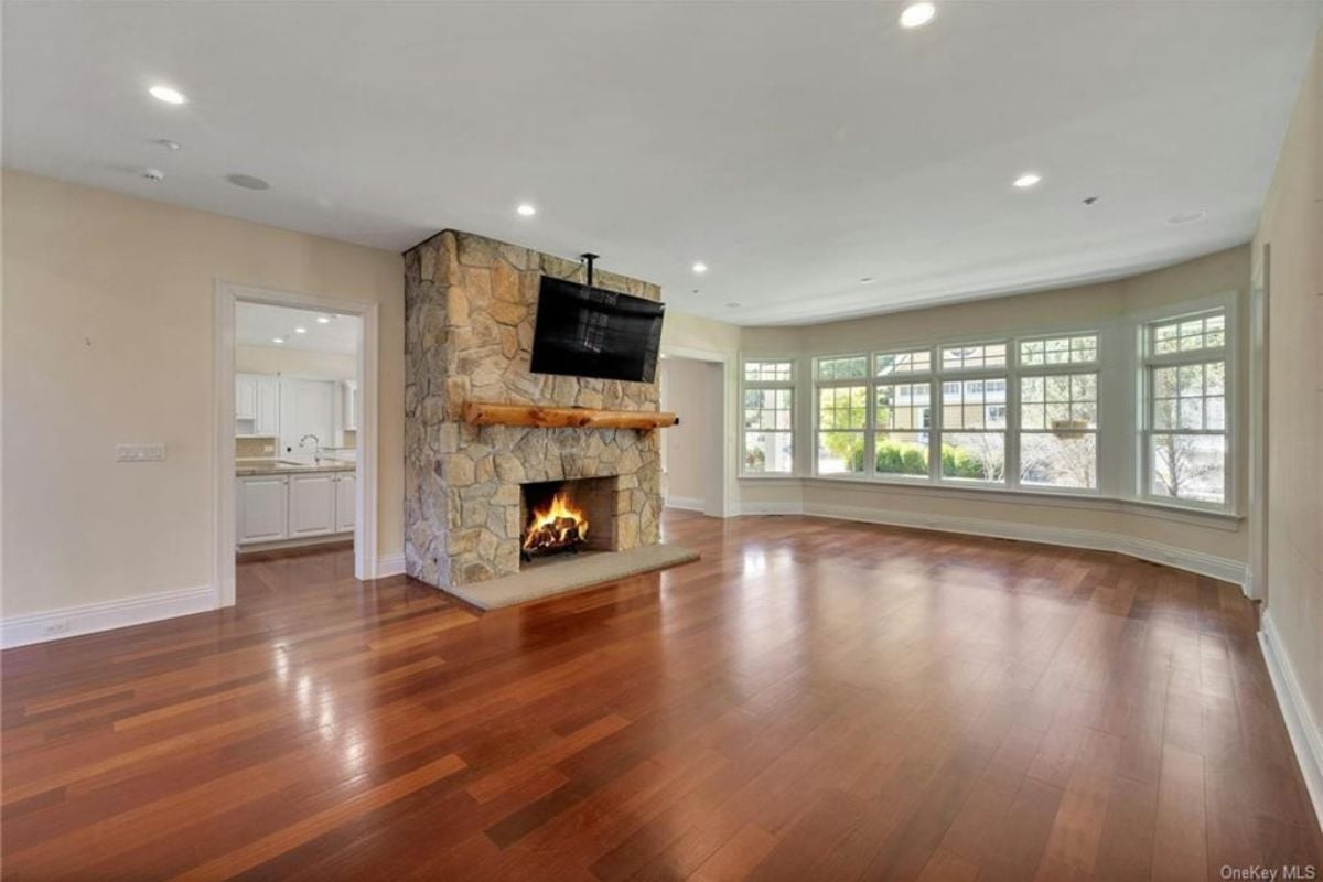 An empty living room with a fireplace, a TV, wooden floors, and windows.