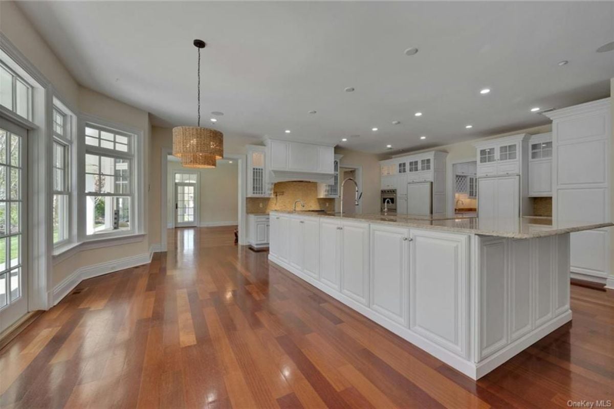 A kitchen with windows, a wooden floor, an island, a white ceiling, and white cabinets.