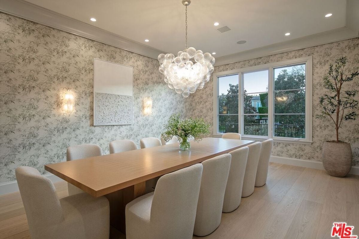 A dining room with wallpaper, chandelier, window, houseplant, and dining table surrounded by chairs.