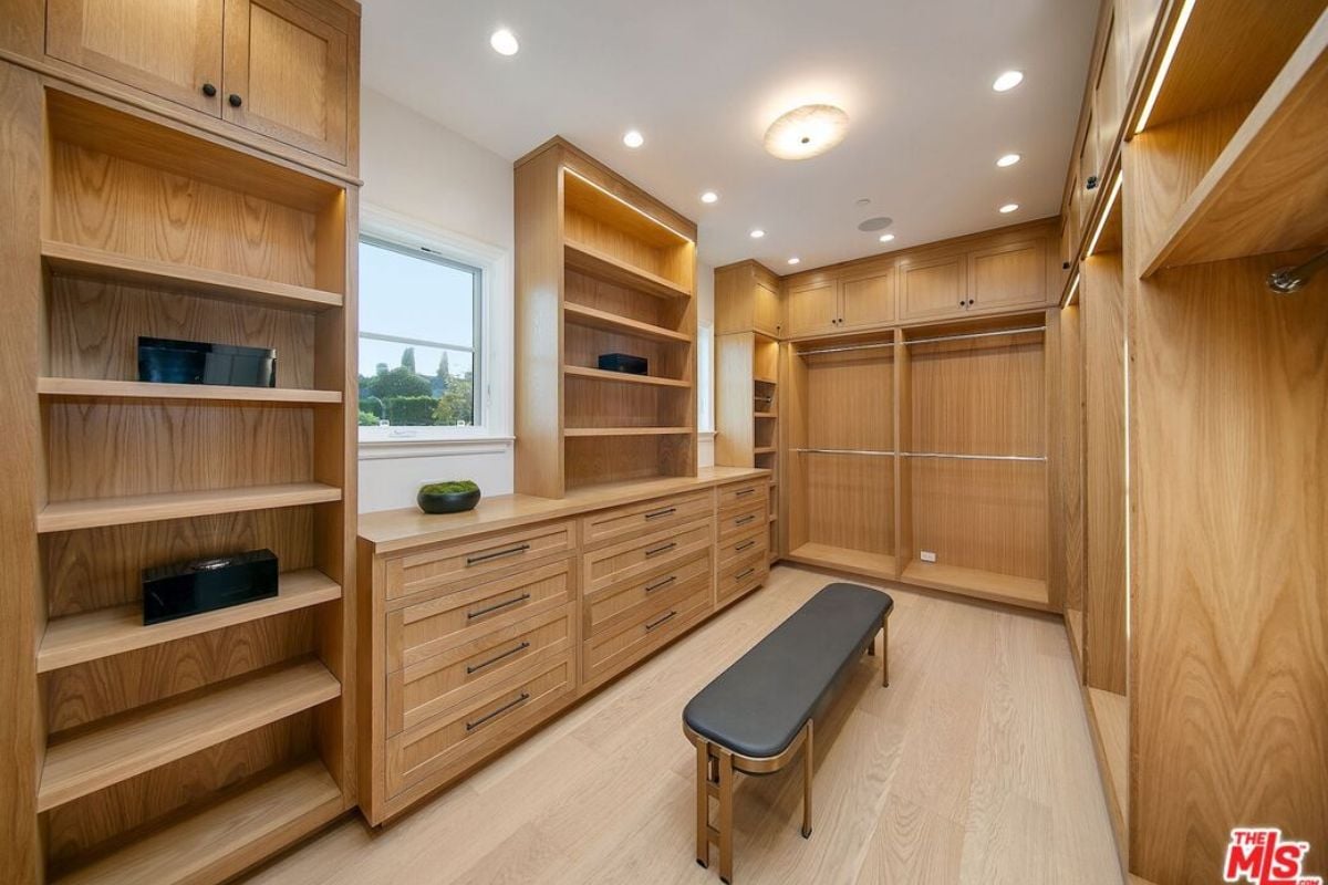 A walk-in closet with a wooden floor, bench, wooden shelves, and cabinets.