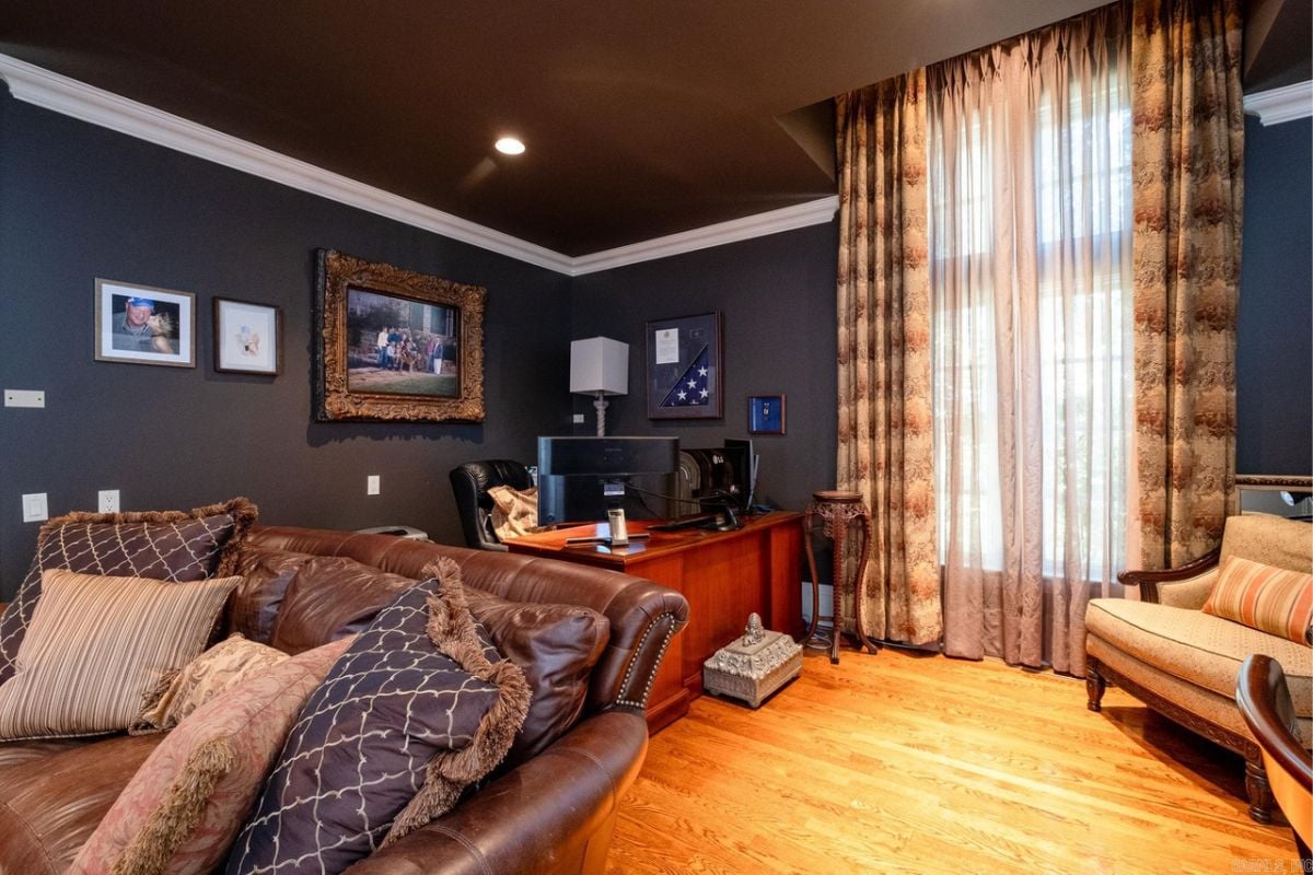 A home office with paintings, couches with pillows, a window with curtains, desk, and chair.