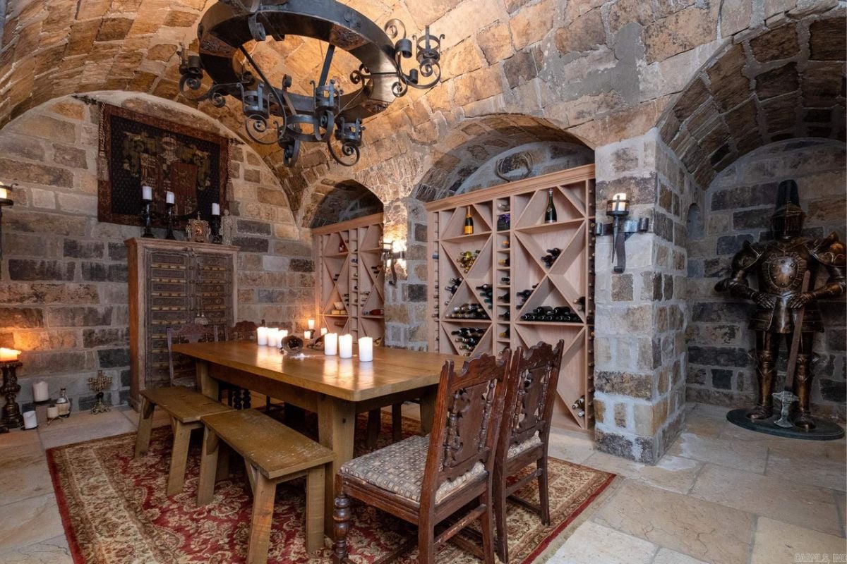 A wine cellar with brick walls and ceiling, wine rack filled with wine bottles, and a table surrounded by chairs and benches.