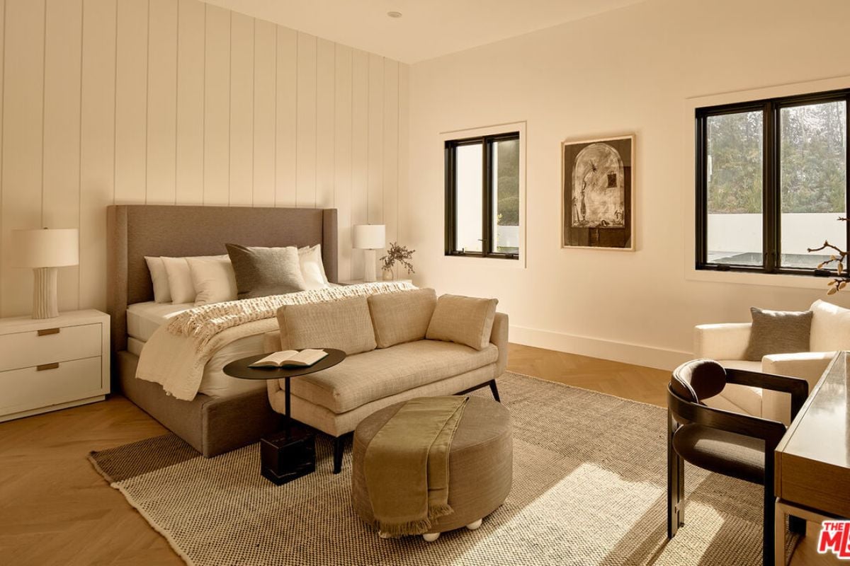 A bedroom with a bed, rug, nightstand, lampshade, couch, chair, and windows.