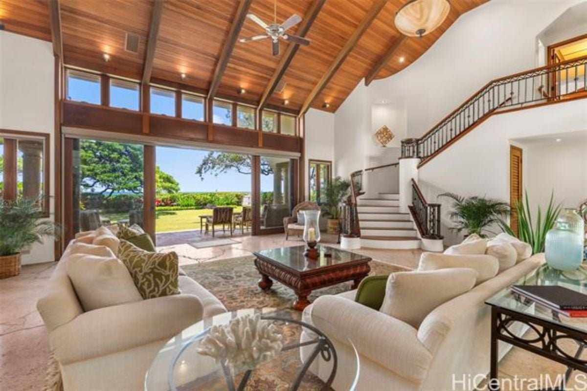 A living room with an exposed beam ceiling, ceiling fan, house plants, rug, white couches, and a center table.