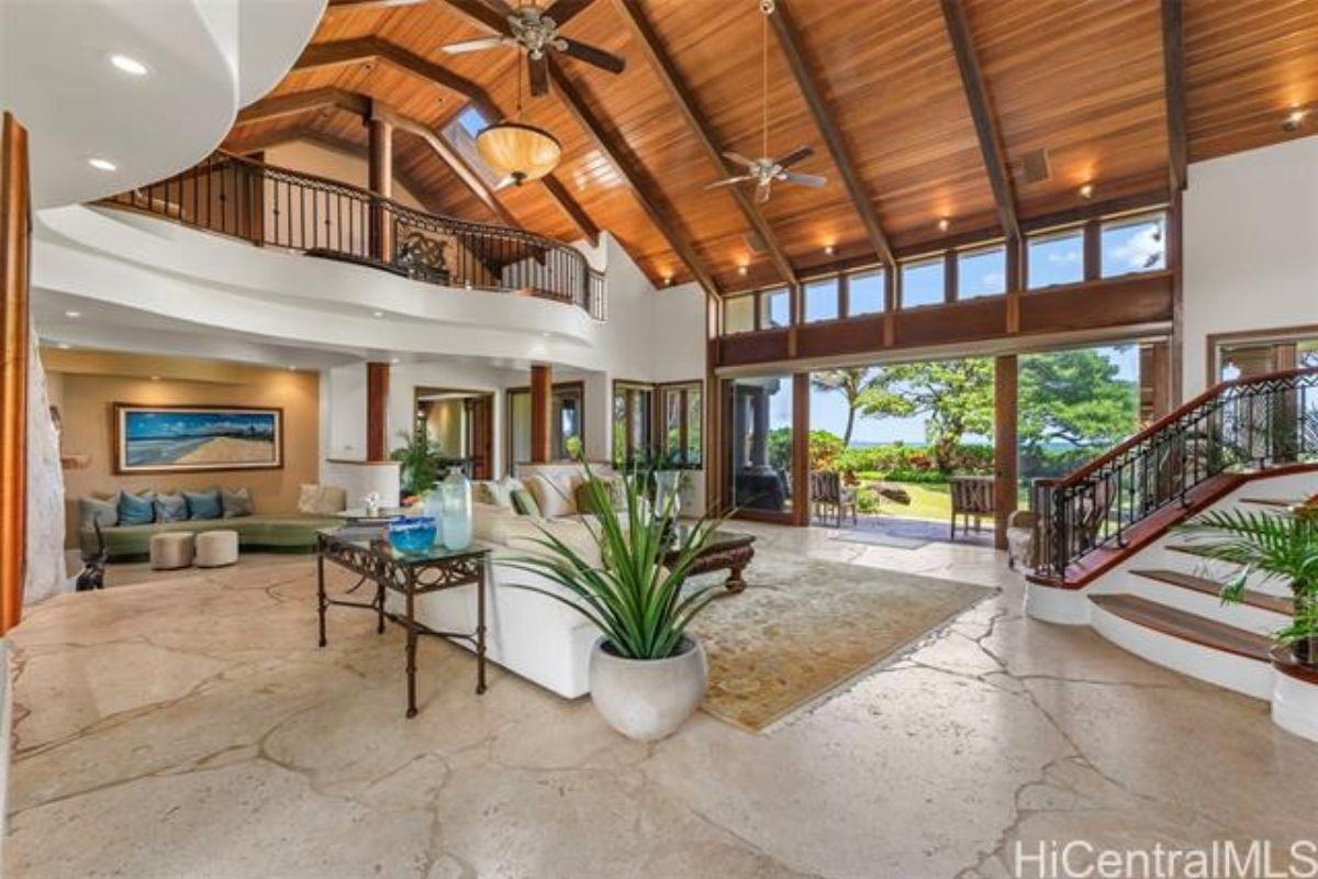 A living room with an exposed beam ceiling, ceiling fan, house plants, rug, white couches, and a center table.