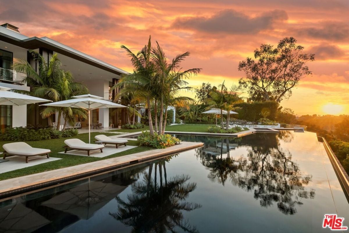 A swimming pool with trees, umbrellas, and chairs on the poolside.