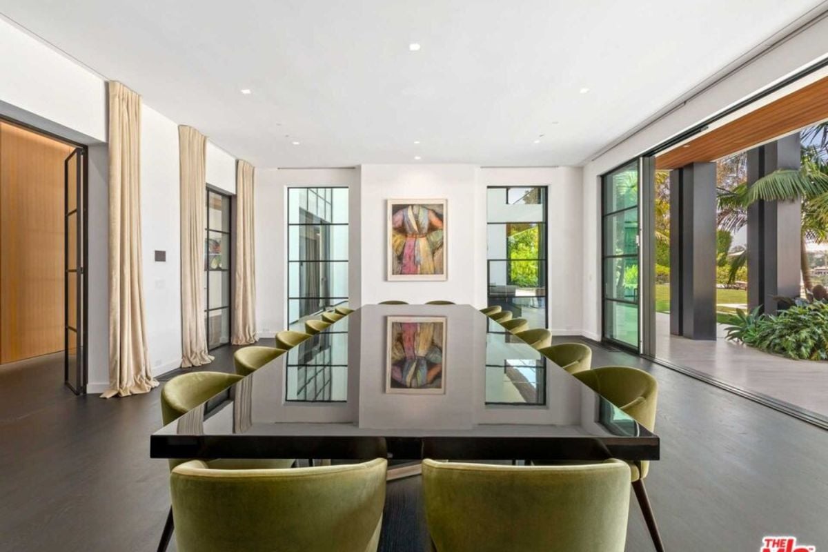 A dining room with a large glass sliding door, windows, a painting, and a long dining table surrounded by green chairs.