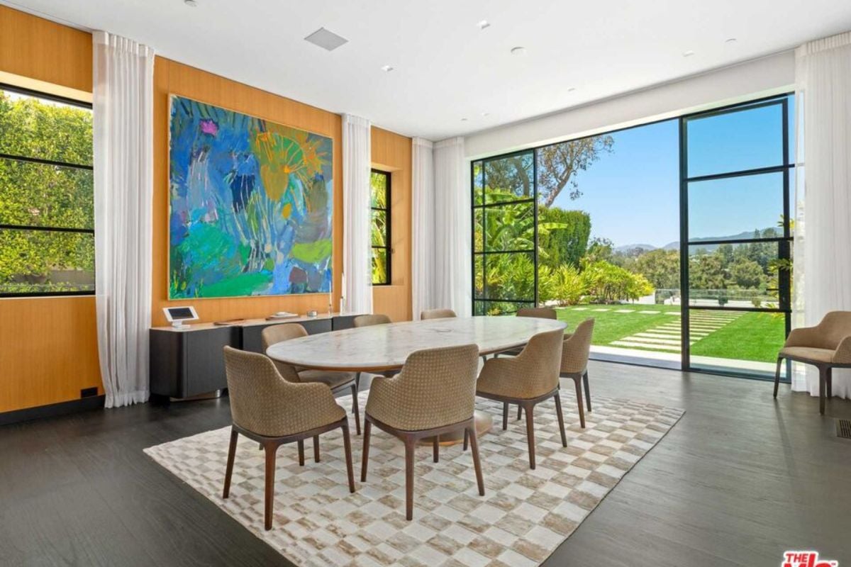A dining room with a rug, painting, windows, and a dining table surrounded by chairs.