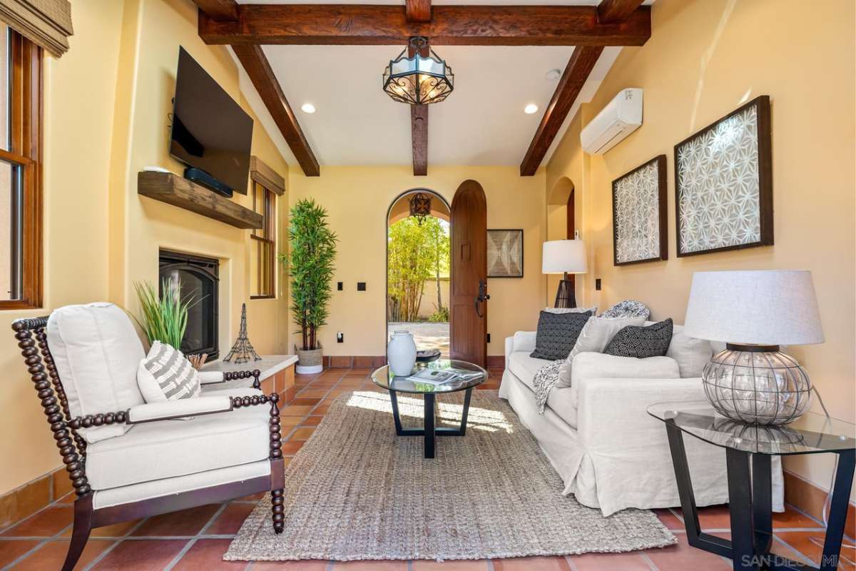 A living room with a beamed ceiling, fireplace, TV, houseplants, couch, chairs, and a coffee table.