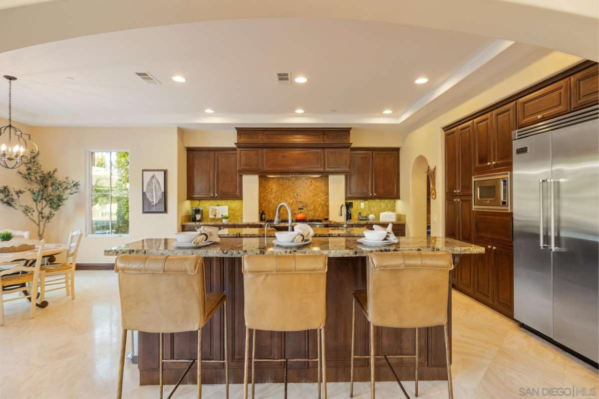 A kitchen with a countertop, stools, sink, wooden cabinets, and kitchen appliances.