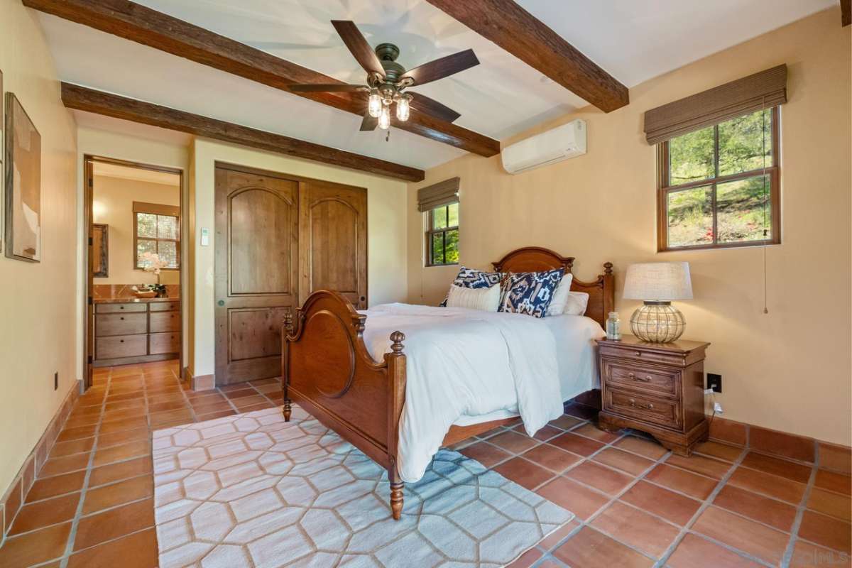 A bedroom with a bed, beamed ceiling, ceiling fan, windows, nightstand, and lampshade.