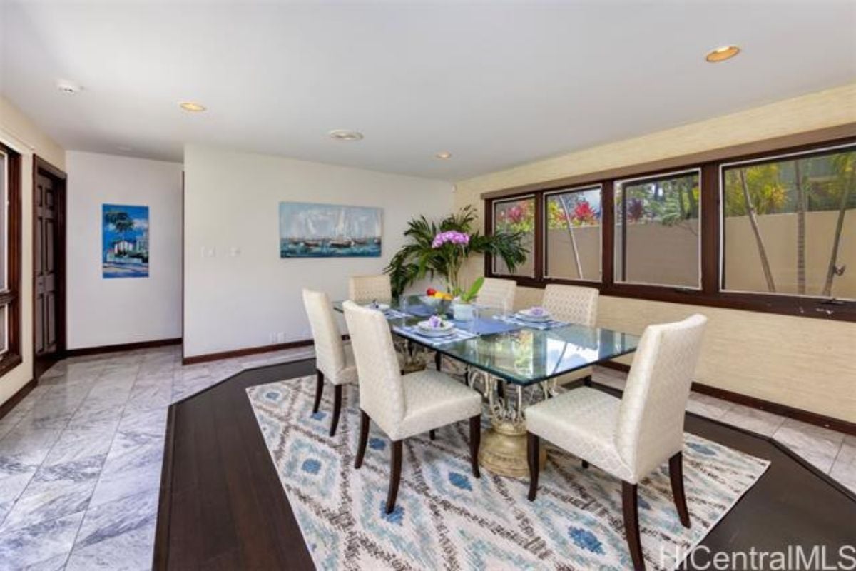 A dining room with windows, paintings, houseplants, and a glass table surrounded by chairs.