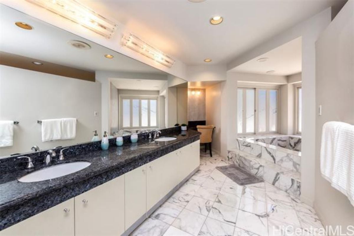 A bathroom with a countertop, mirror, cabinets, chair, tiled floor, windows, and bathtub.