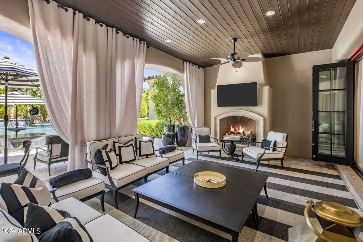A patio with a fireplace, TV, ceiling fan, chairs, couch, and center table.