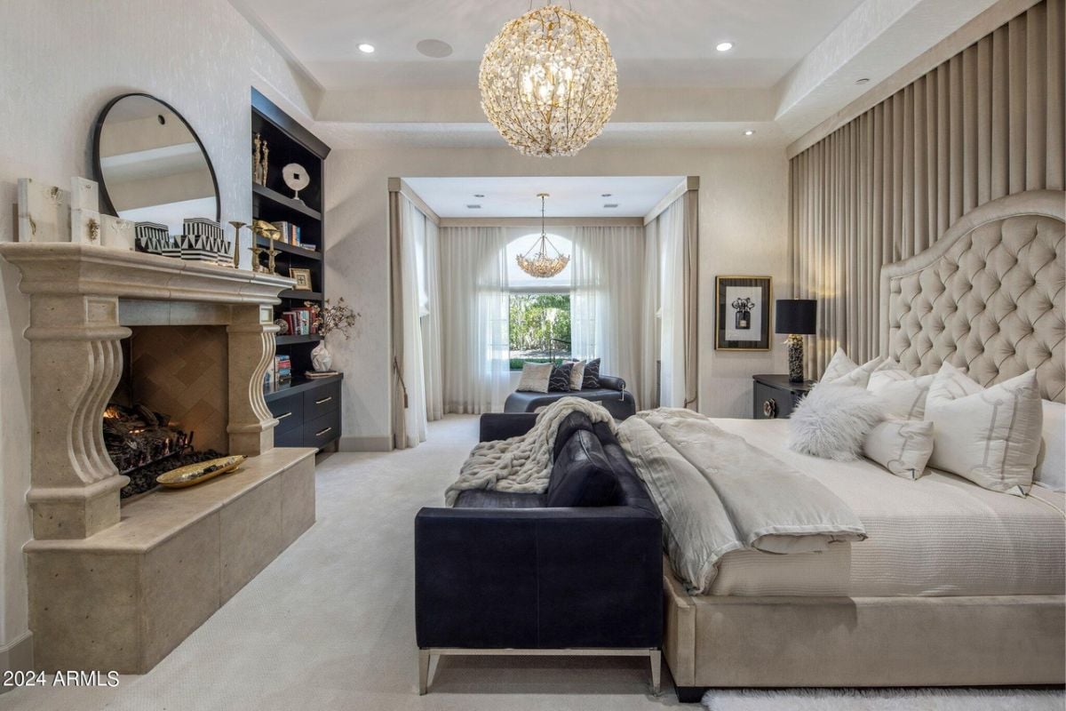 A bedroom with a bed, headboard, fireplace, chandelier, shelf, couch, and window.