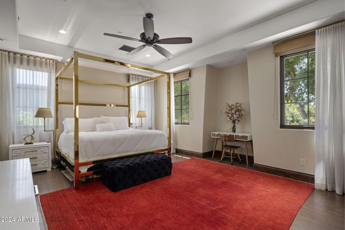 A bedroom with a bed with canopy, nightstand, ceiling fan, table, chair, red rug, and window.