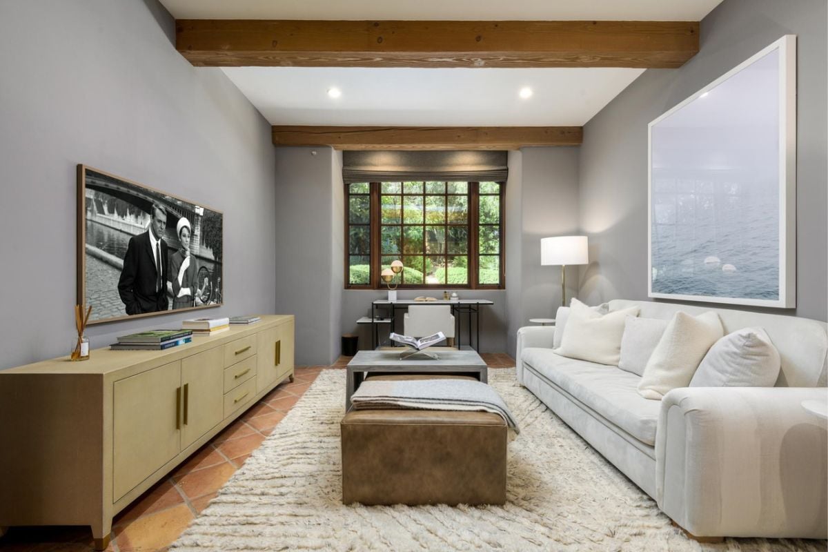 A lounge with a TV, cabinets, a window, a painting, a beamed ceiling, a white couch, and a center table.